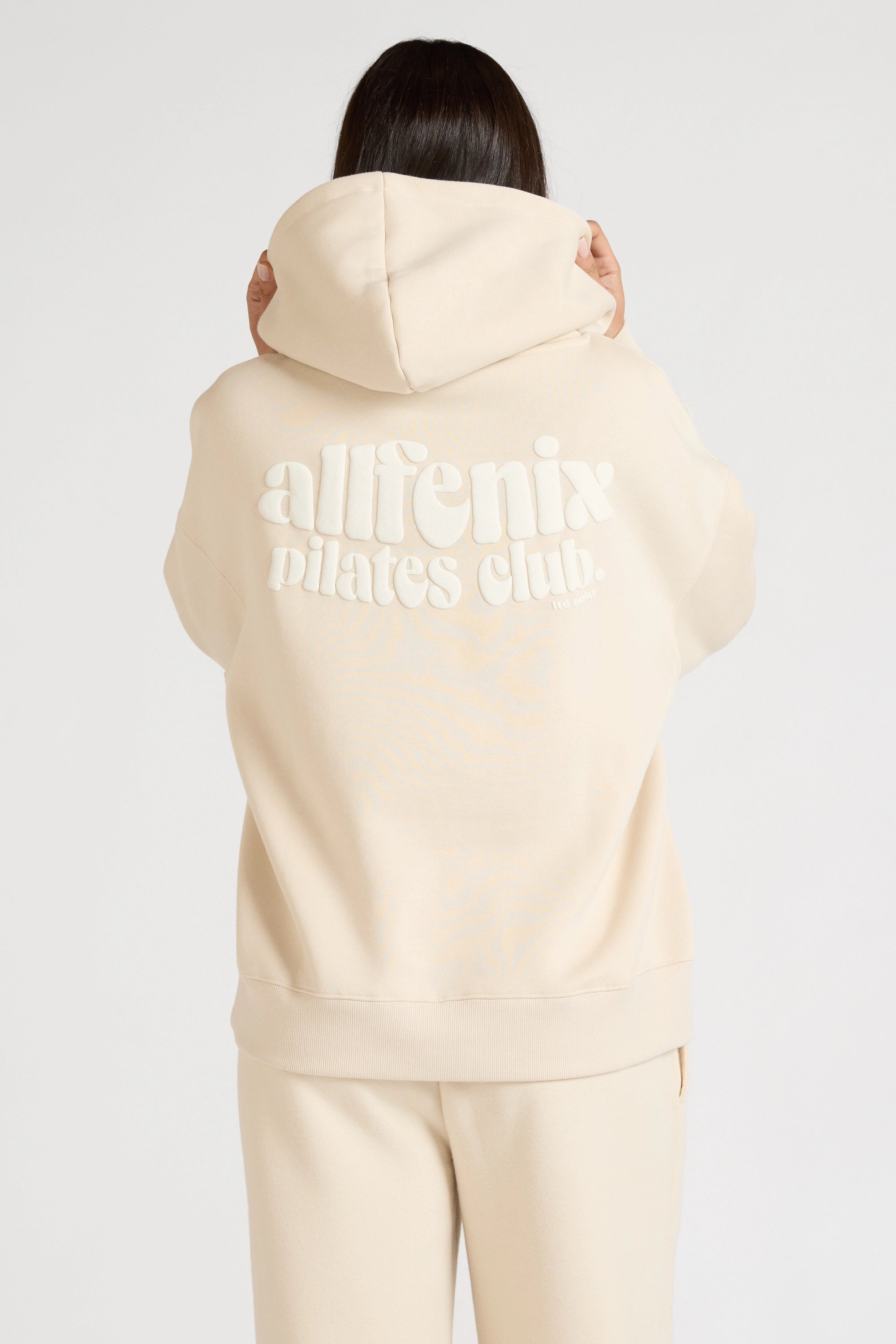 Women wearing a beige hoodie with 'allfenix pilates club' text on the back against a white background