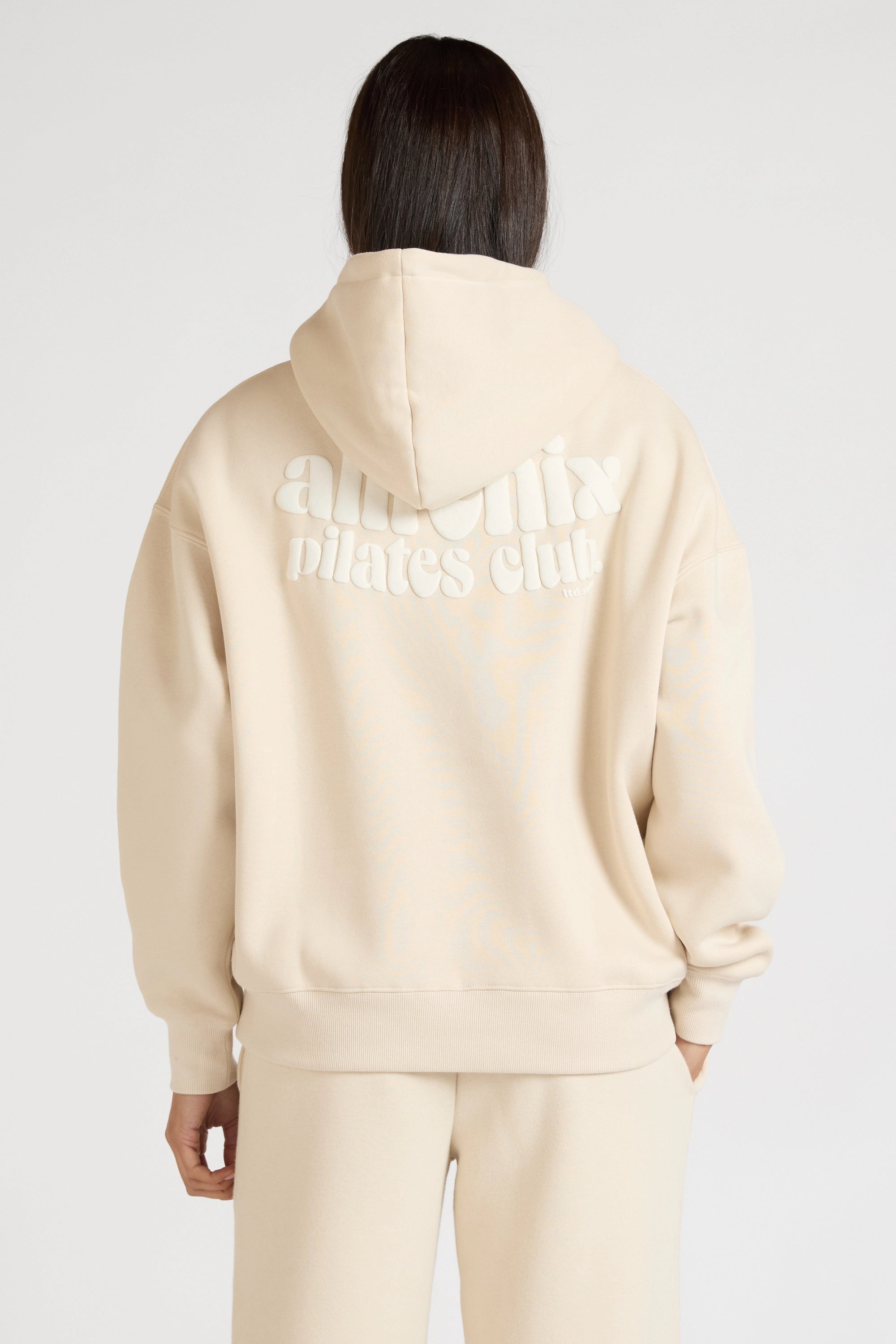 Woman wearing a beige hoodie with 'allfenix pilates club' text on the back against a white background