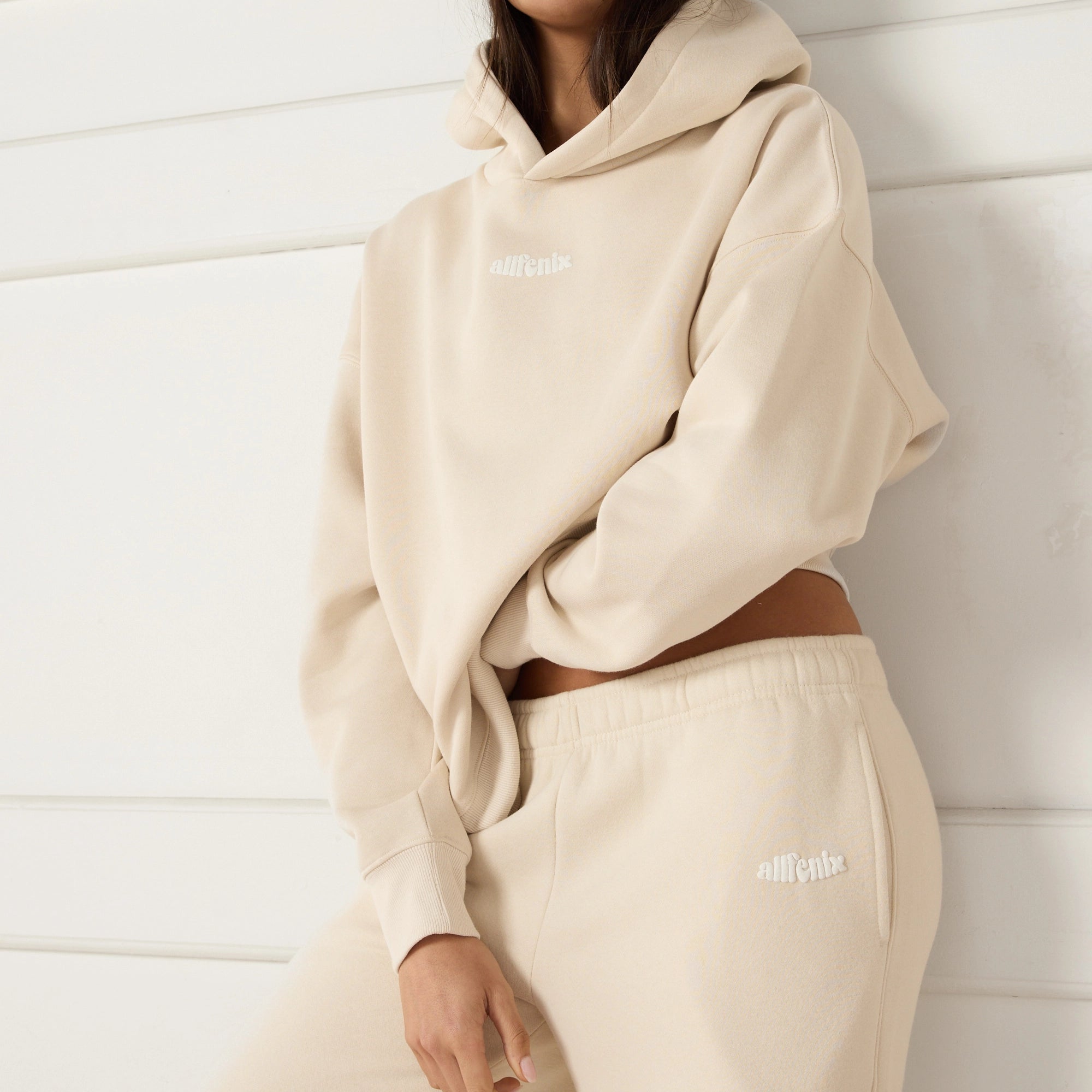 Woman wearing a beige hoodie and pants with a logo against a white background