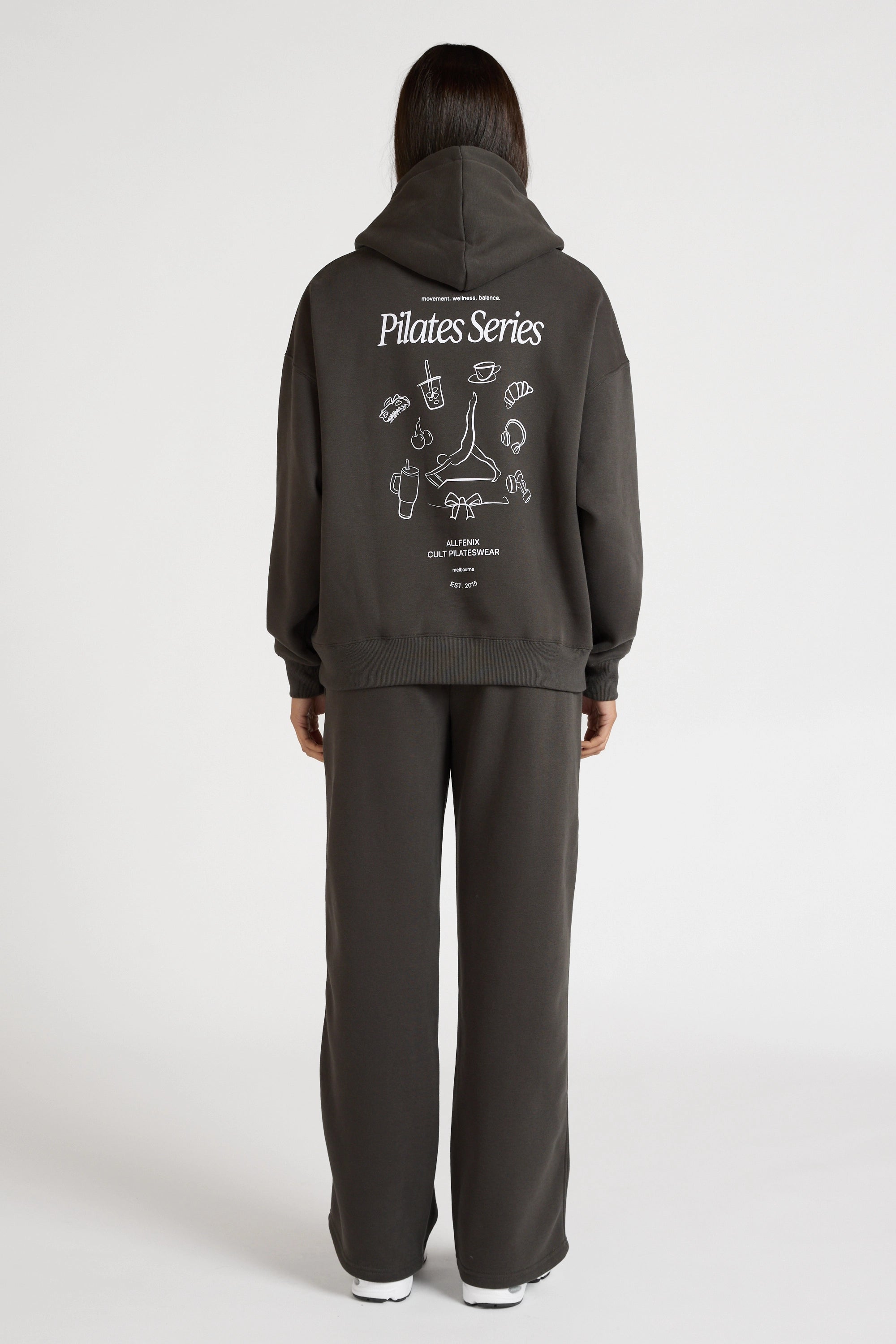 Woman wearing a dark grey hoodie and pants with 'Pilates Series' design on the back, standing against a white background.