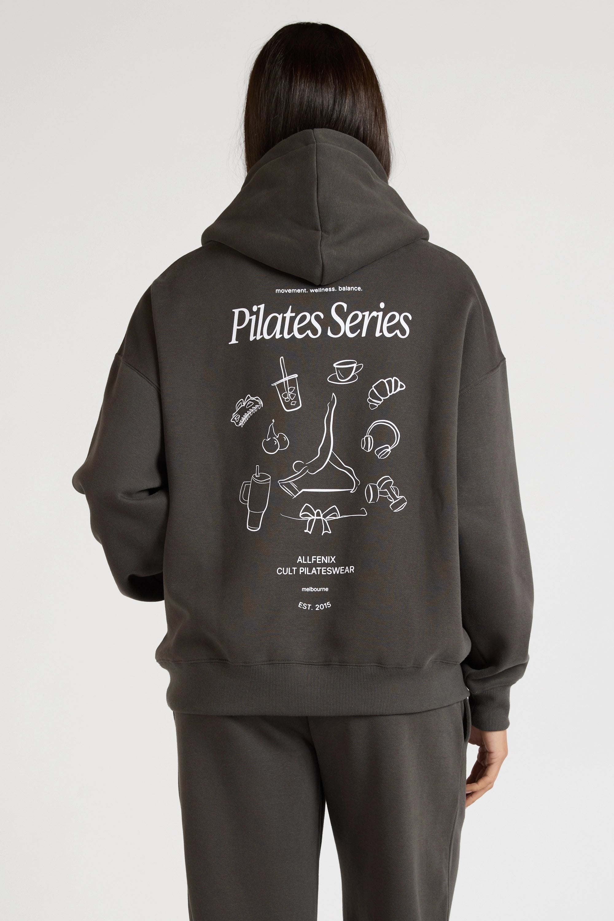 Woman wearing a dark grey hoodie with 'Pilates Series' design on the back against a white background