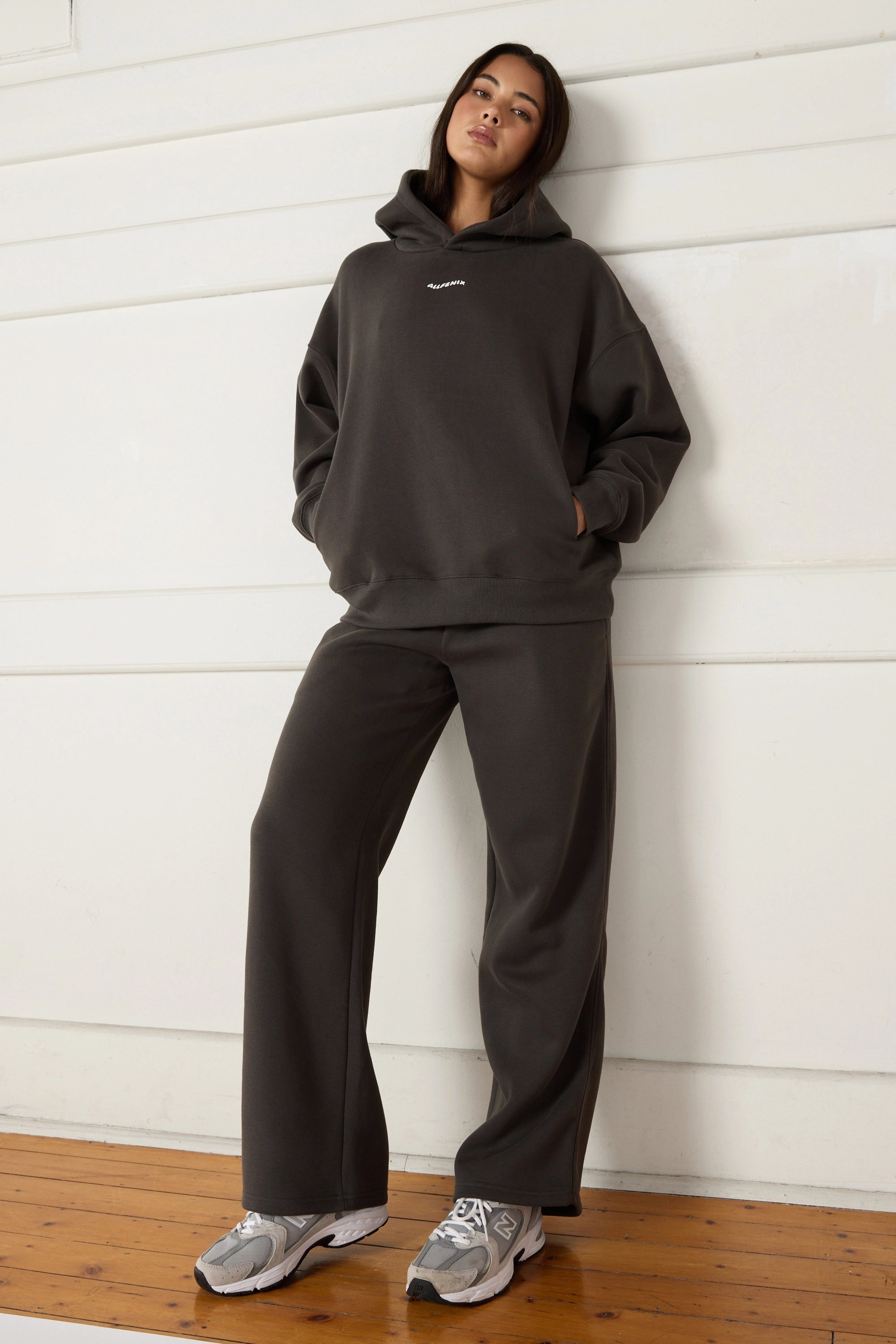 Woman wearing a dark grey hoodie and pants leaning against a white wall.