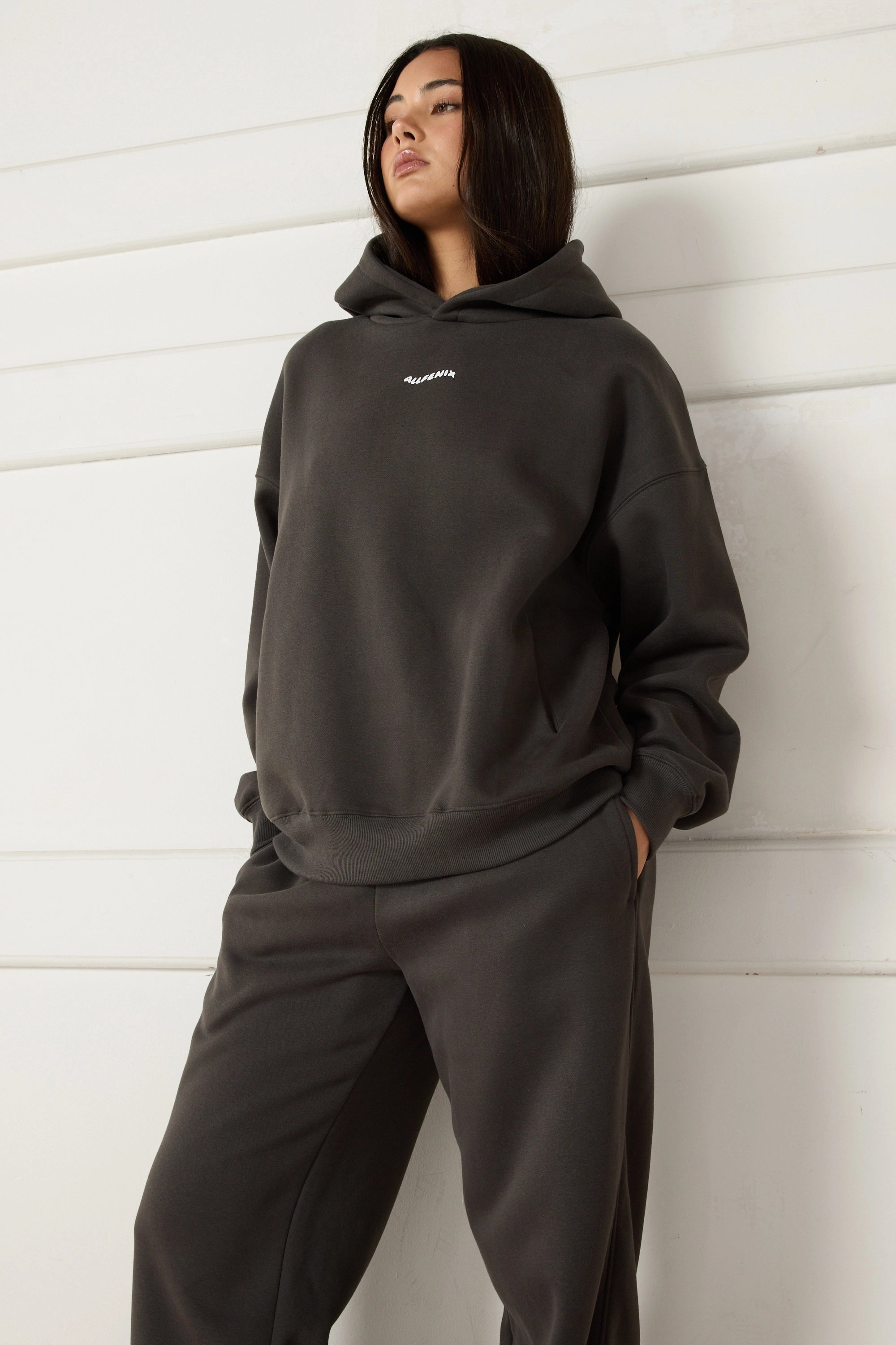 Woman wearing a dark grey hoodie and pants against a white wall