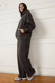 Woman wearing a dark grey hoodie and pants standing against a white wall.