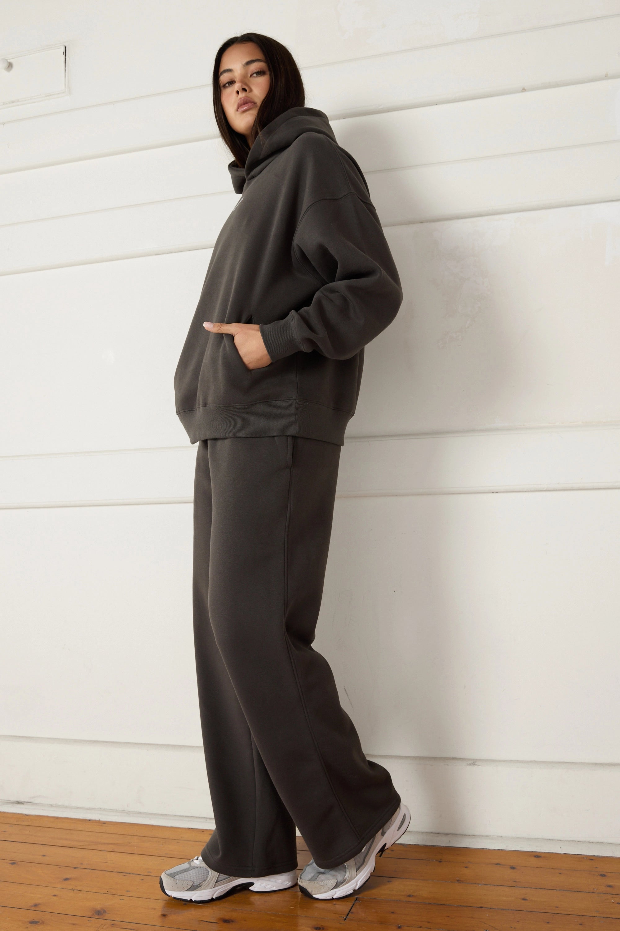 Woman wearing a dark grey hoodie and pants standing against a white wall.