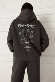 Woman wearing a dark grey hoodie with 'Pilates Series' design on the back against a white wall.