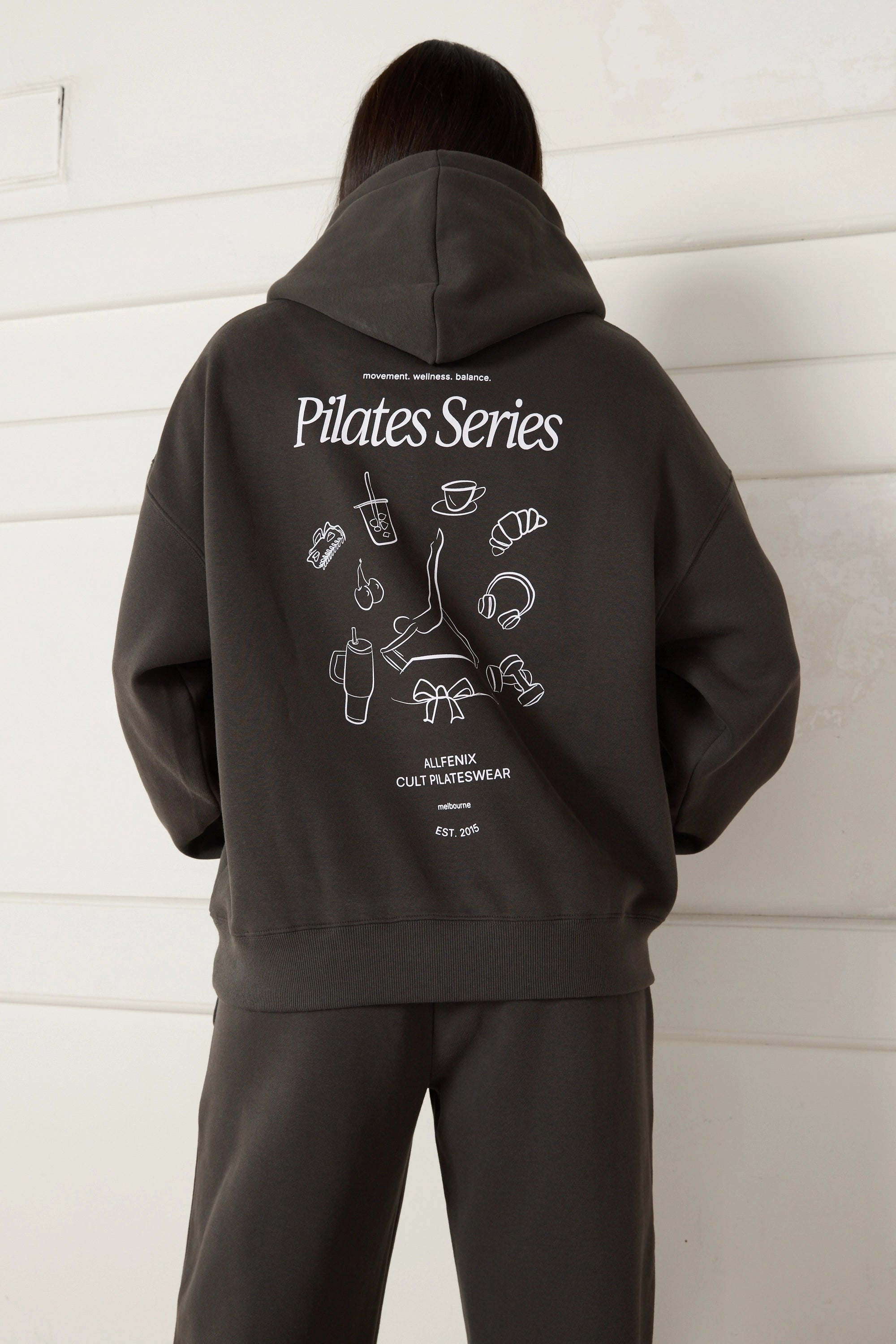 Woman wearing a dark grey hoodie with 'Pilates Series' design on the back against a white wall.