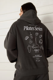 Woman wearing a dark grey hoodie with 'Pilates Series' design on the back against a white wall.