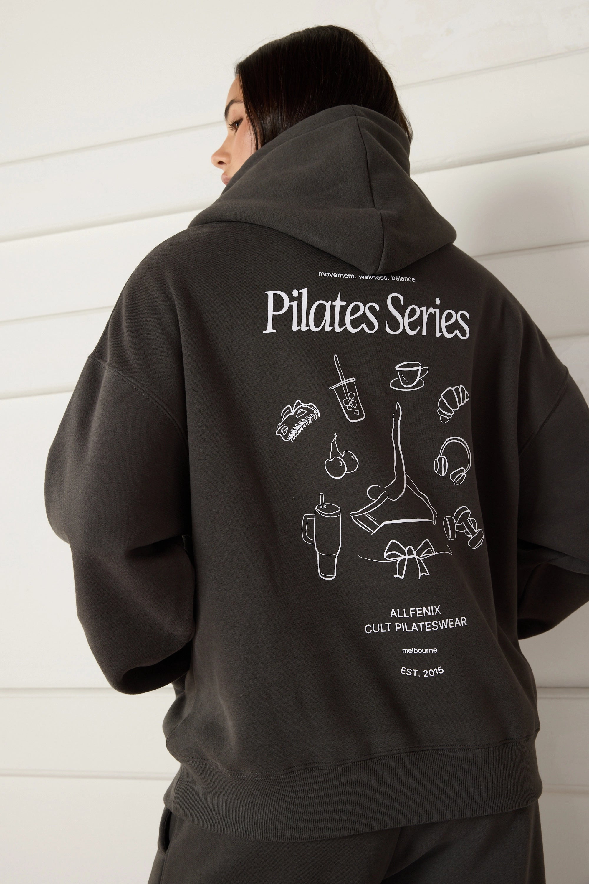 Woman wearing a dark grey hoodie with 'Pilates Series' design on the back against a white wall.