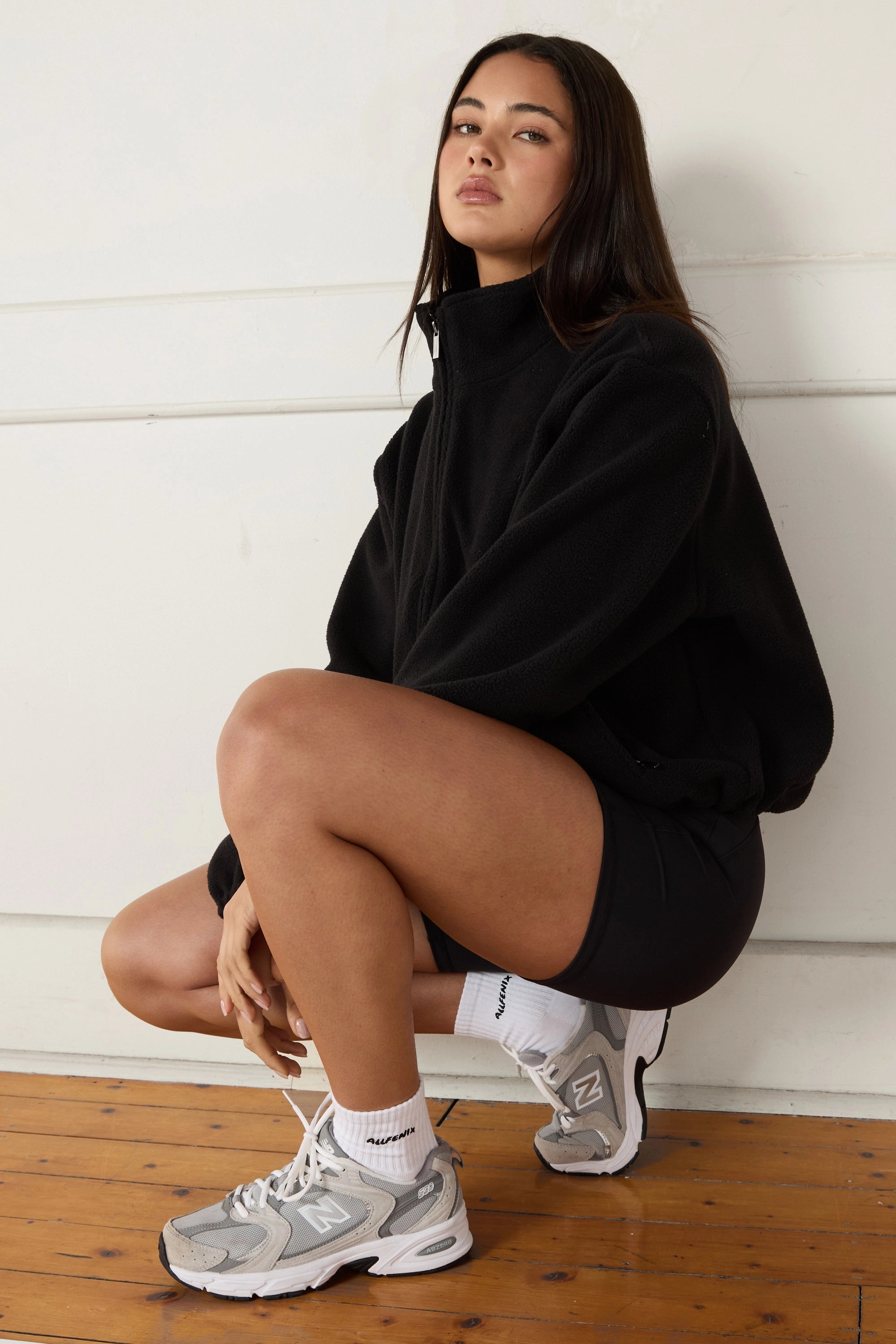 Woman in black soft fleece jacket with white socks and grey sneakers sitting on a wooden floor.