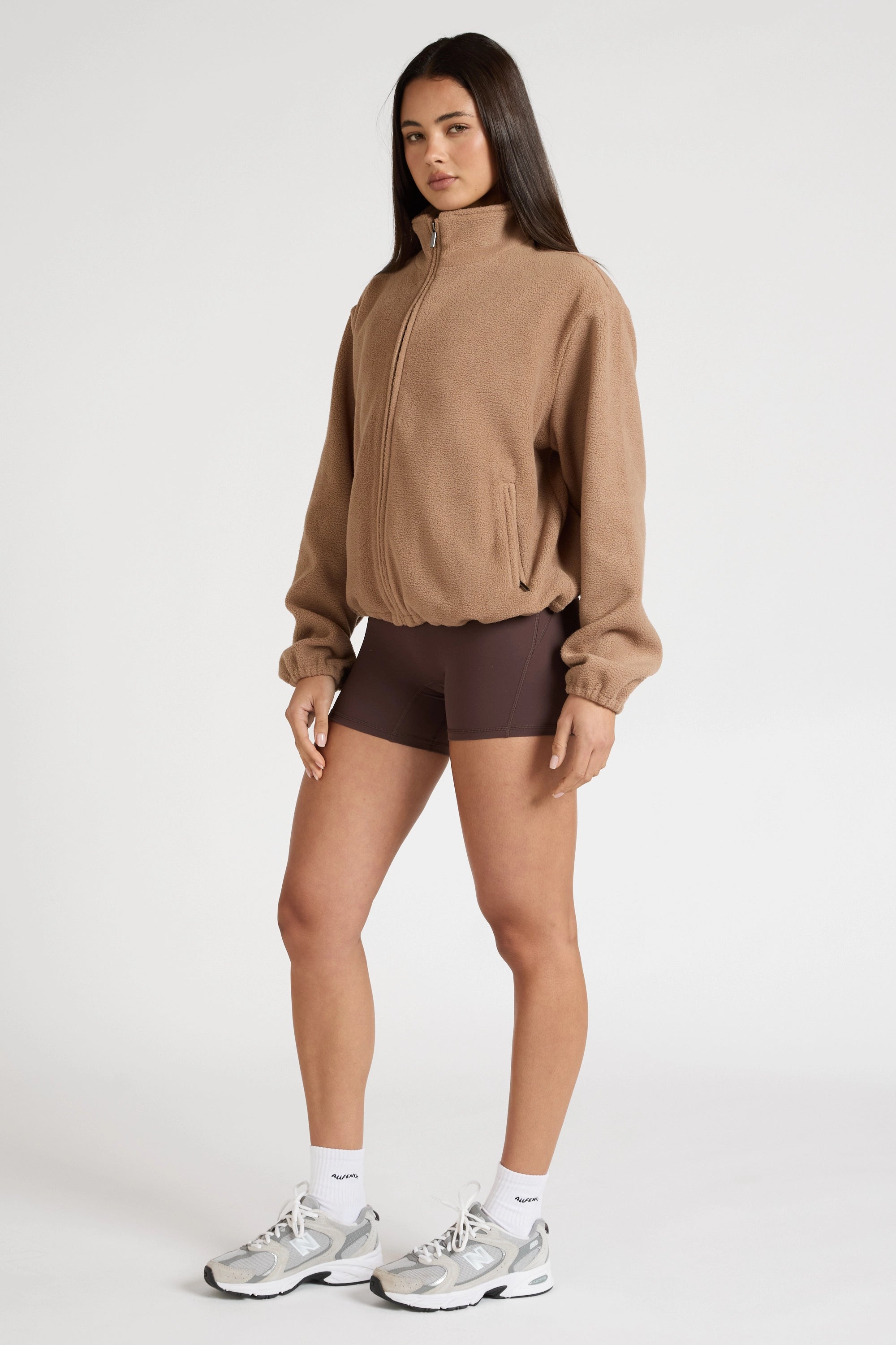 Woman wearing a brown soft fleece zip jacket and shorts with white sneakers on a white background